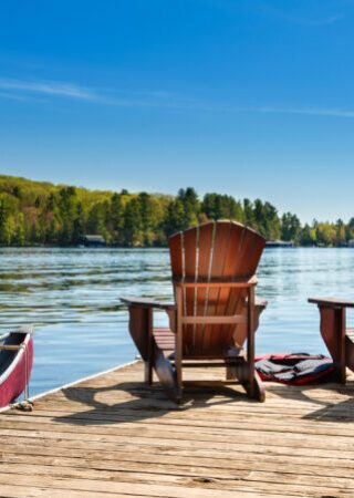 Muskoka chairs on a wooden dock Two Muskoka chairs on a wooden dock overlooking the blue water of a lake in Muskoka, Ontario Canada. A red canoe is tied to the pier and life jackets are visible near the chairs.